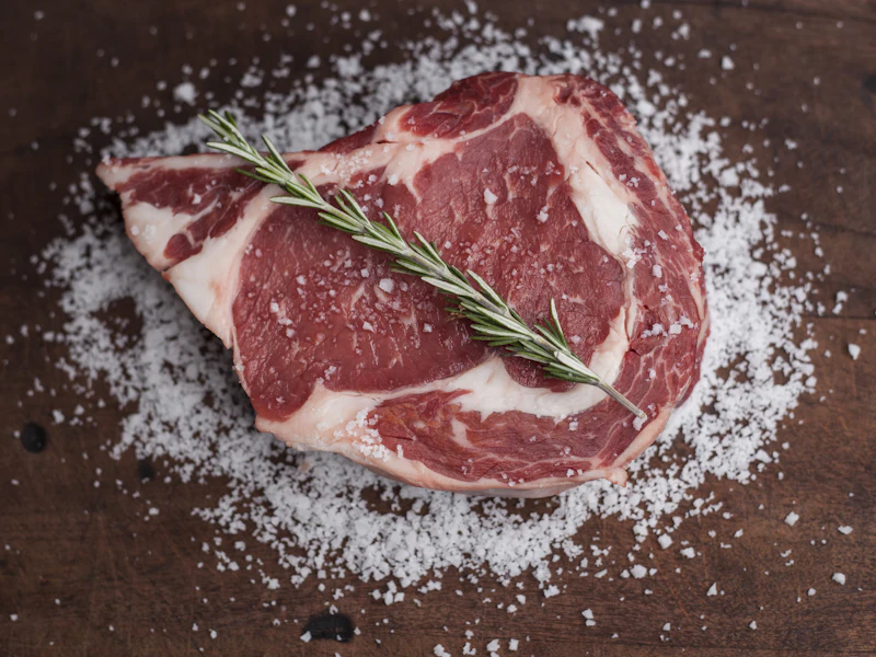 Rohes Rib-Eye auf grobem Salz, daneben ein Zweig Rosmarin.