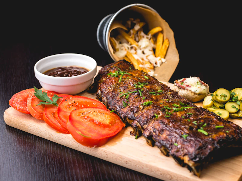 Short Ribs auf einem Holzbrett, daneben Tomaten und eine Schale Sauce.